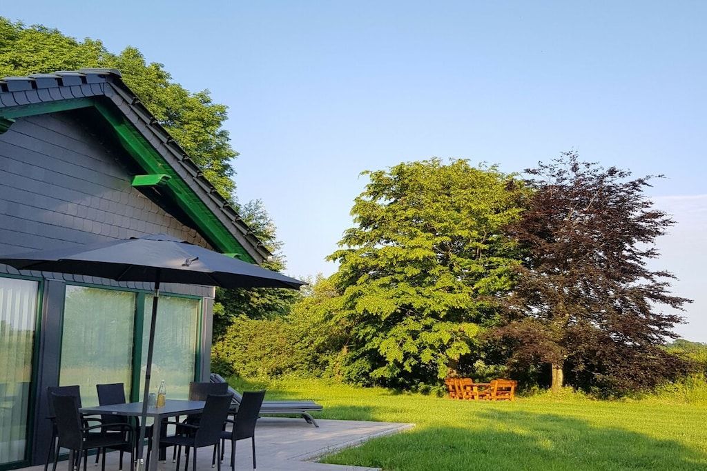 A modern building with a covered patio area, outdoor seating, and a large tree in the background.
