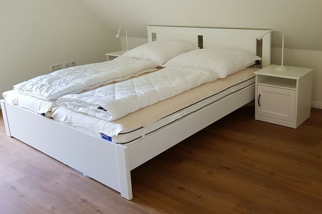 Een eenpersoonskamer met een wit hoofdeinde, een nachtkastje met een lamp en een klein kastje.