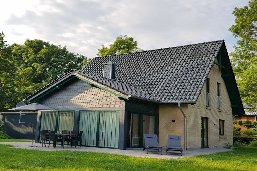 Une maison à deux étages, avec un toit en tuiles, une terrasse couverte avec des chaises et une pelouse.
