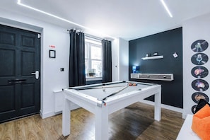Una mesa de billar en una habitación con una puerta negra, una ventana con cortinas, un estante con objetos y una pared con discos de vinilo.