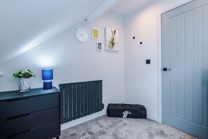 A room with a radiator, a clock, a lamp, a door, and a bag on the floor. - Stylish Home for 8 | Games Room & Free Parking (Northwich)