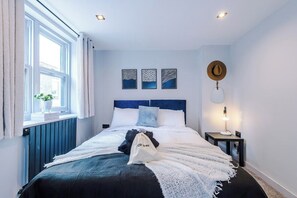 A bedroom with a bed, a bedside table with a lamp, a window with curtains, and wall art.
