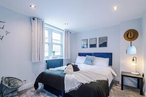 A bedroom with a bed, a bedside table with a lamp, a window with curtains, and wall art. - Stylish Home for 8 | Games Room & Free Parking (Northwich)