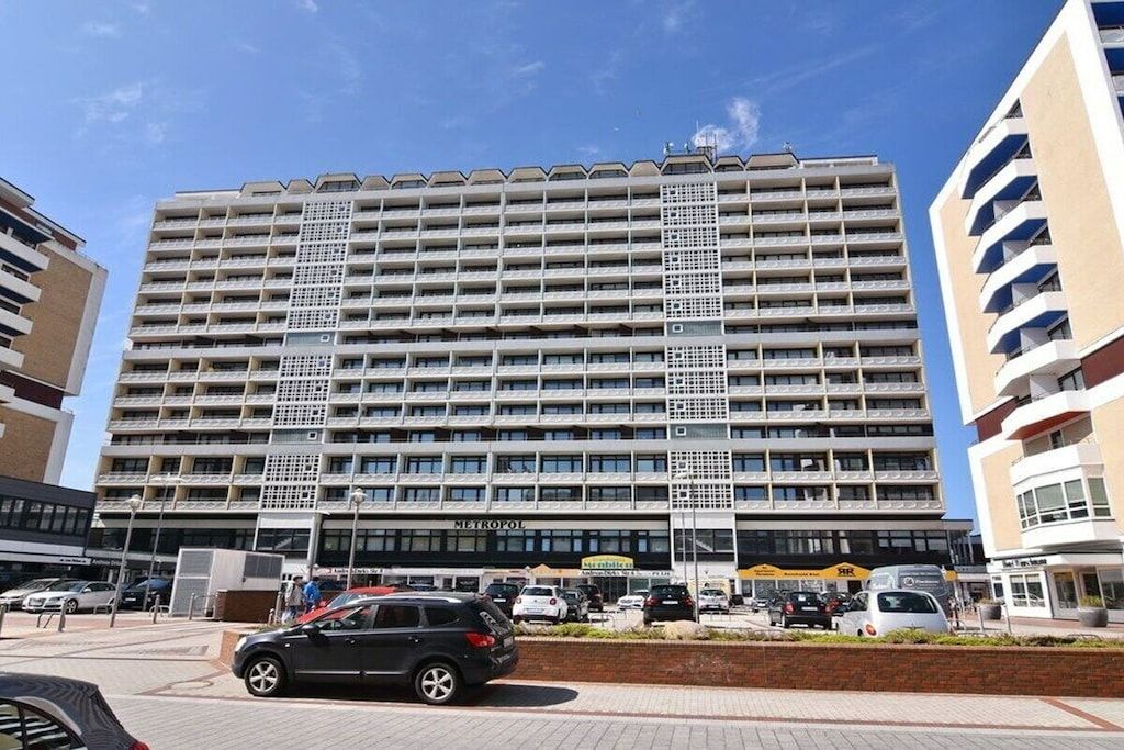 A multi-story hotel with a central entrance, a parking area in front, and a clear sky.