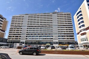 Ein mehrstöckiges Hotel mit zentralem Eingang, einem Parkplatz davor und blauem Himmel.