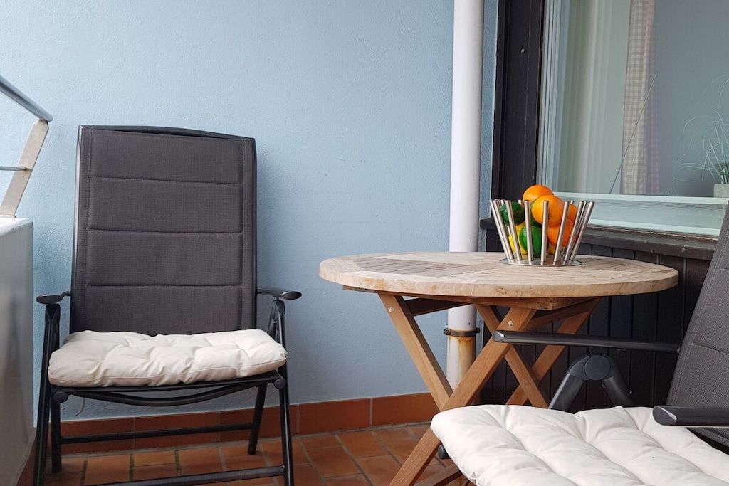 A balcony with a chair, a small round table with a cushion, and a window with a curtain.