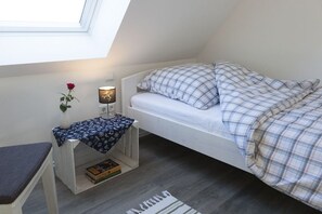 Ein kleiner, gemütlicher Raum mit schrägem Dach, einem Einzelbett mit Karo-Bettwäsche, einem Nachttisch mit Lampe, einem kleinen Stuhl und einem Fenster mit Aussicht.
