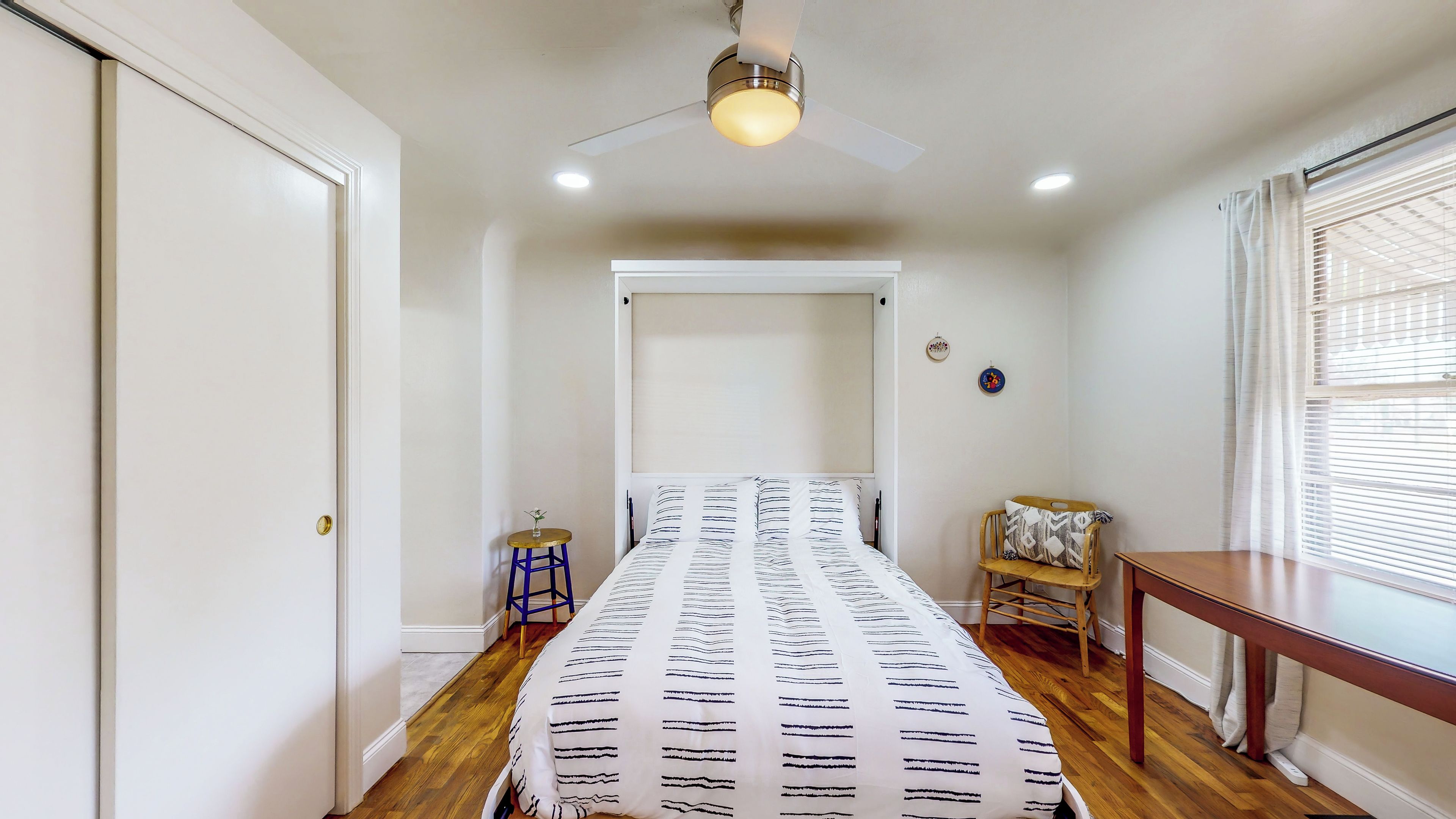 A bedroom with a bed, a ceiling fan, a window with blinds, a small table, and a chair.