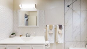 Un baño con un lavabo blanco, un espejo, un perchero con toallas y una zona de ducha.