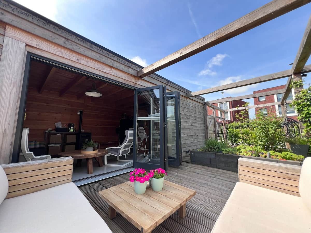 Een terras buiten met houten meubels, een tafel met bloemen en een glazen deur naar een binnenruimte.
