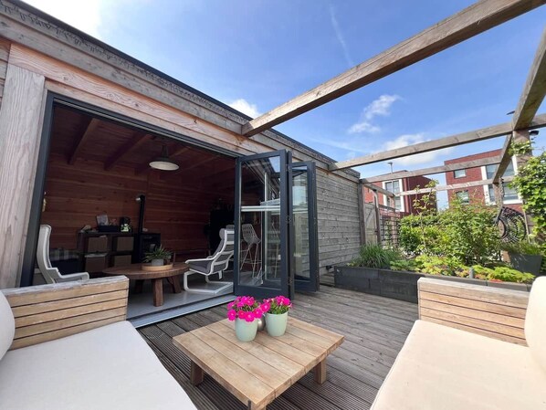 Een terras buiten met houten meubels, een tafel met bloemen en een glazen deur naar een binnenruimte.