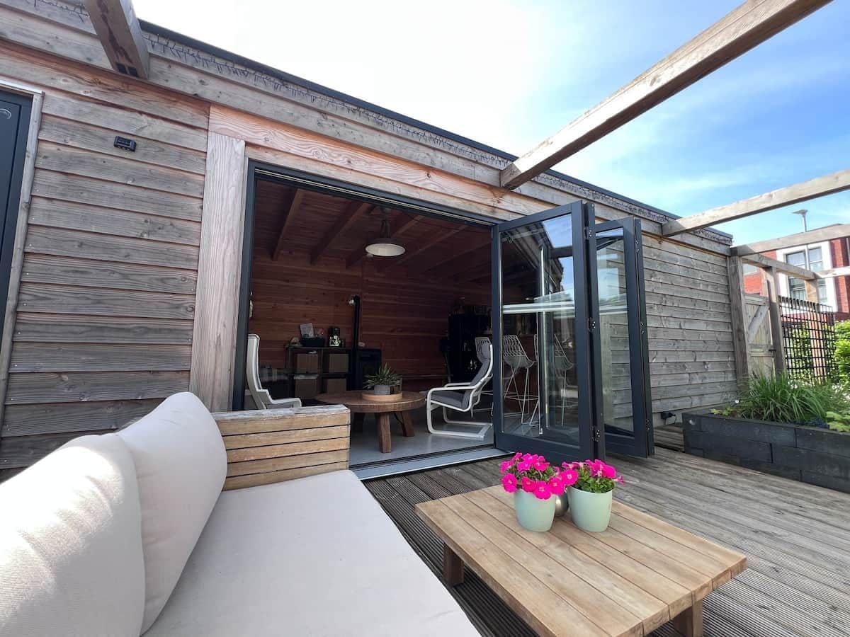 Een houten terras met een bank, een tafel met bloemen en een glazen deur die leidt naar een binnenruimte met stoelen en een tafel.