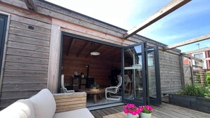 Une terrasse en bois avec un canapé, une table décorée de fleurs et une porte vitrée donnant sur un espace intérieur aménagé avec des chaises et une table.