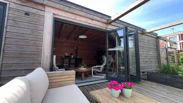Een houten terras met een bank, een tafel met bloemen en een glazen deur die leidt naar een binnenruimte met stoelen en een tafel.