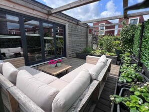 Un espacio al aire libre con sofás y una mesa, rodeado de plantas y una terraza de madera.