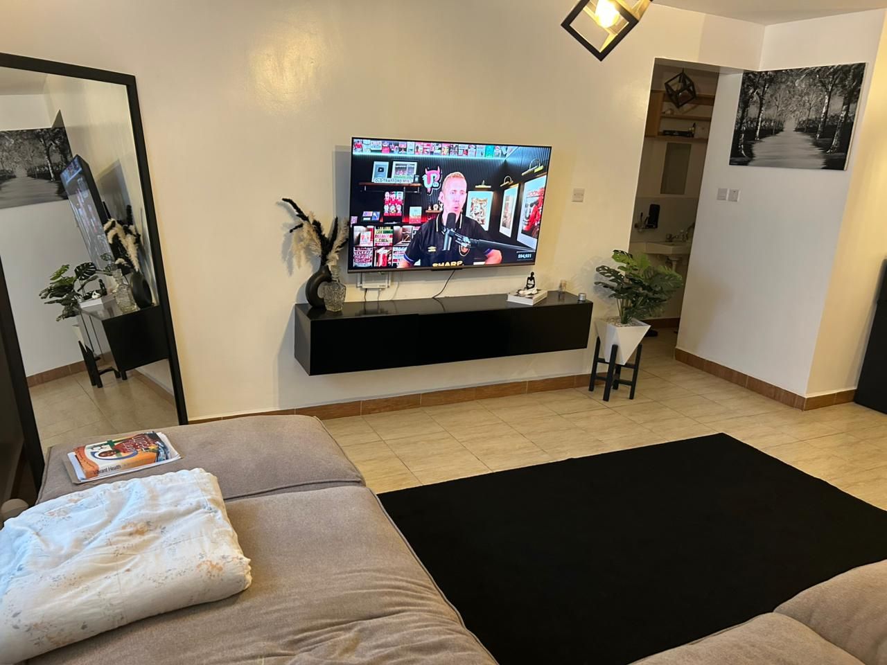 Een woonkamer met een flatscreen televisie bevestigd aan een zwart dressoir, een beige bank, een zwart tapijt en een potplant.