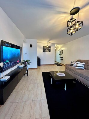 Uma sala de estar moderna com uma TV de tela plana, uma mesa de centro preta, um sofá bege e uma luminária pendente geométrica singular.