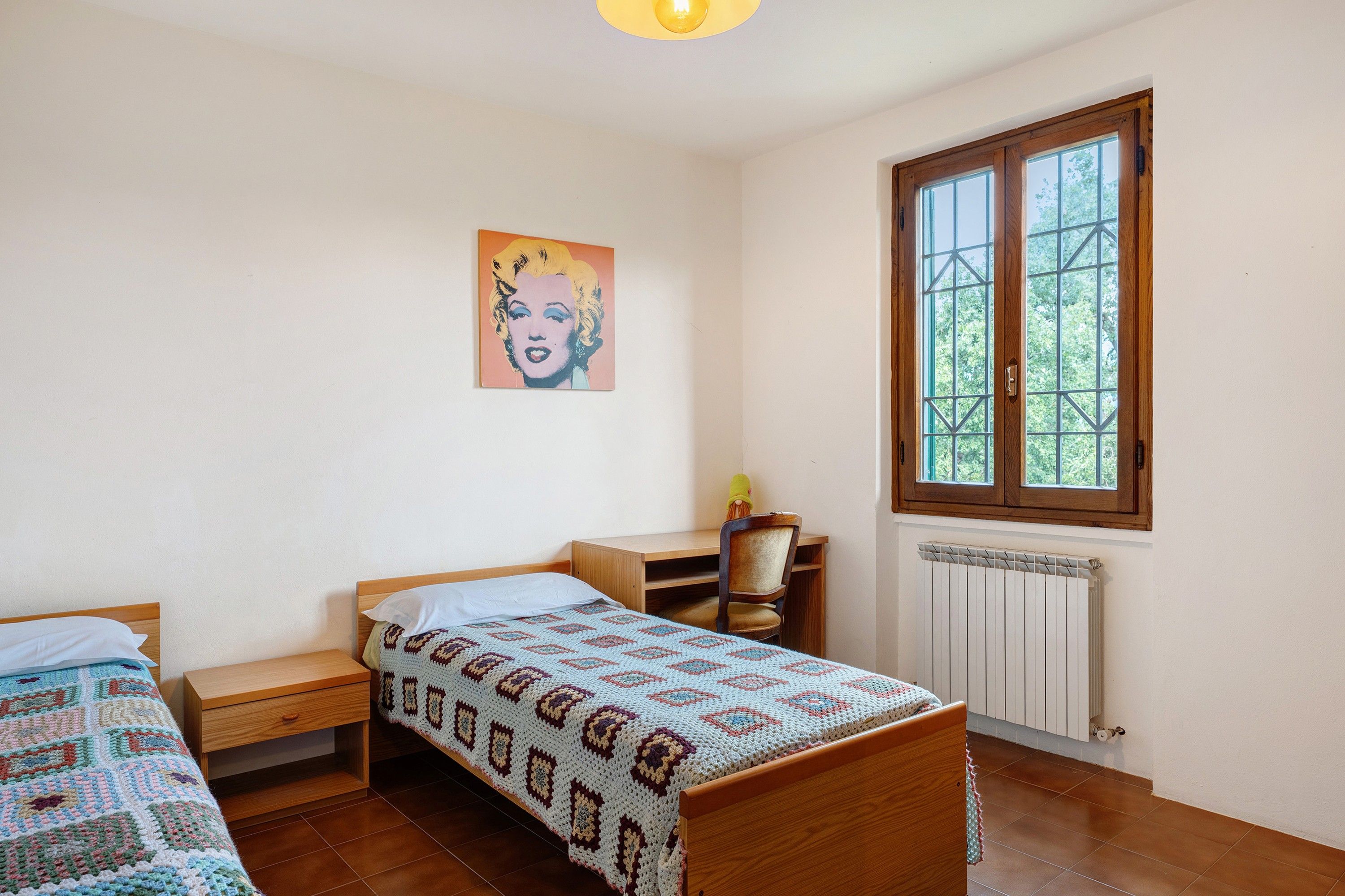 Une chambre avec deux lits simples, un bureau, une chaise, un radiateur et une fenêtre donnant sur des arbres.