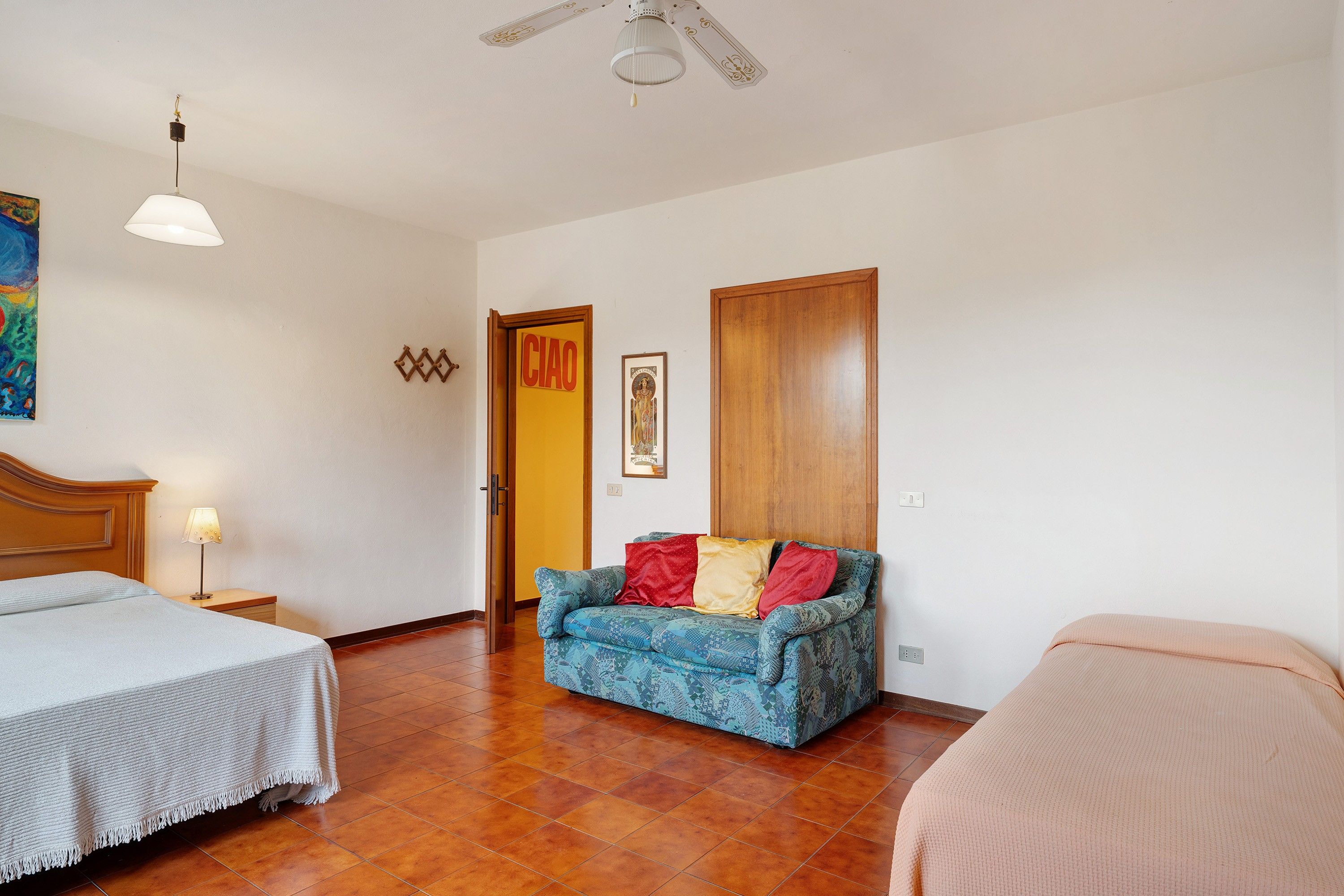 A hotel room with two beds, a sofa, a ceiling fan, a painting, and a door marked 'CIAO'.