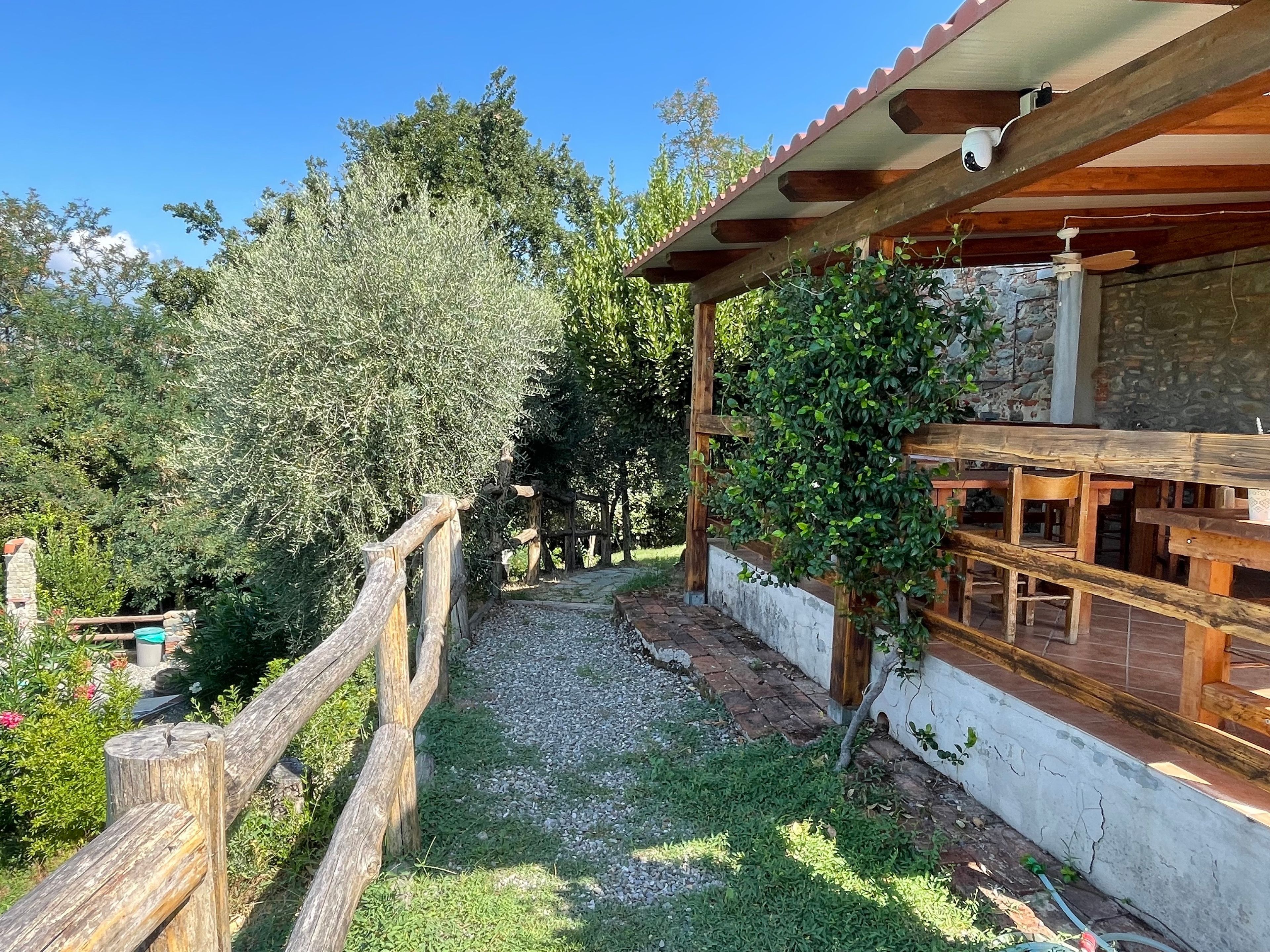 Una pergola in legno con un muro in pietra e un vialetto sterrato che conduce a un'area relax con panche di legno.