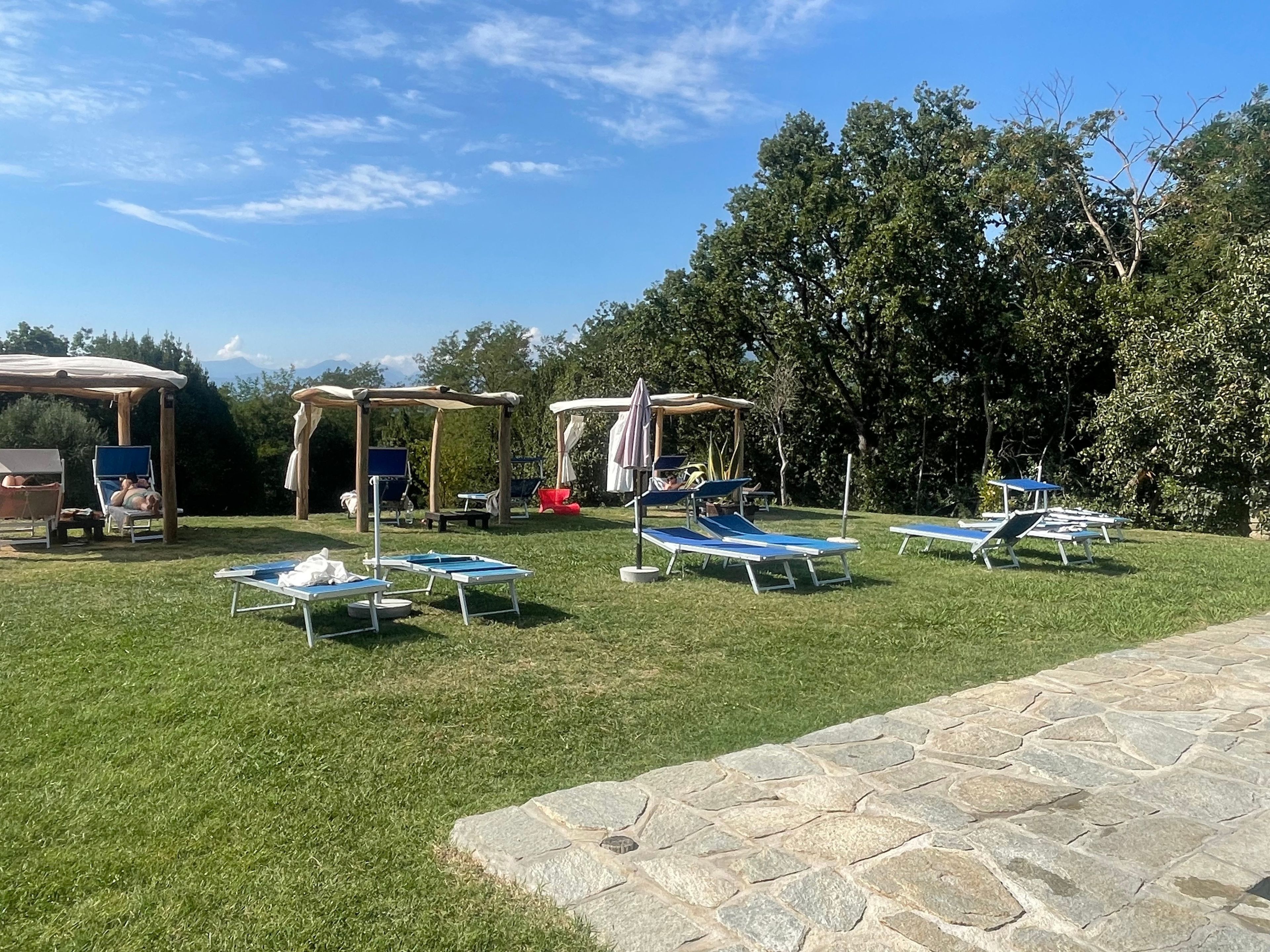 An outdoor area with multiple sun loungers and a covered seating area, surrounded by trees and a stone pathway.