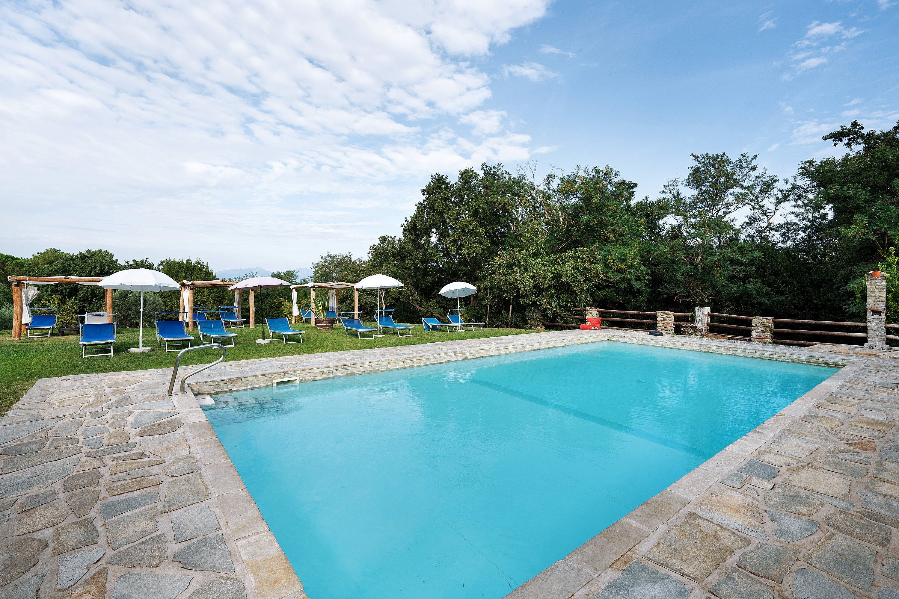 A rectangular swimming pool with stone edging, surrounded by a stone deck. There are blue lounge chairs and white umbrellas.