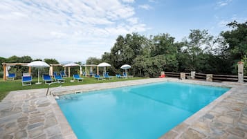 A rectangular swimming pool with stone edging, surrounded by a stone deck. There are blue lounge chairs and white umbrellas.