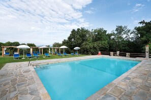 A rectangular swimming pool with stone edging, surrounded by a stone deck. There are blue lounge chairs and white umbrellas.