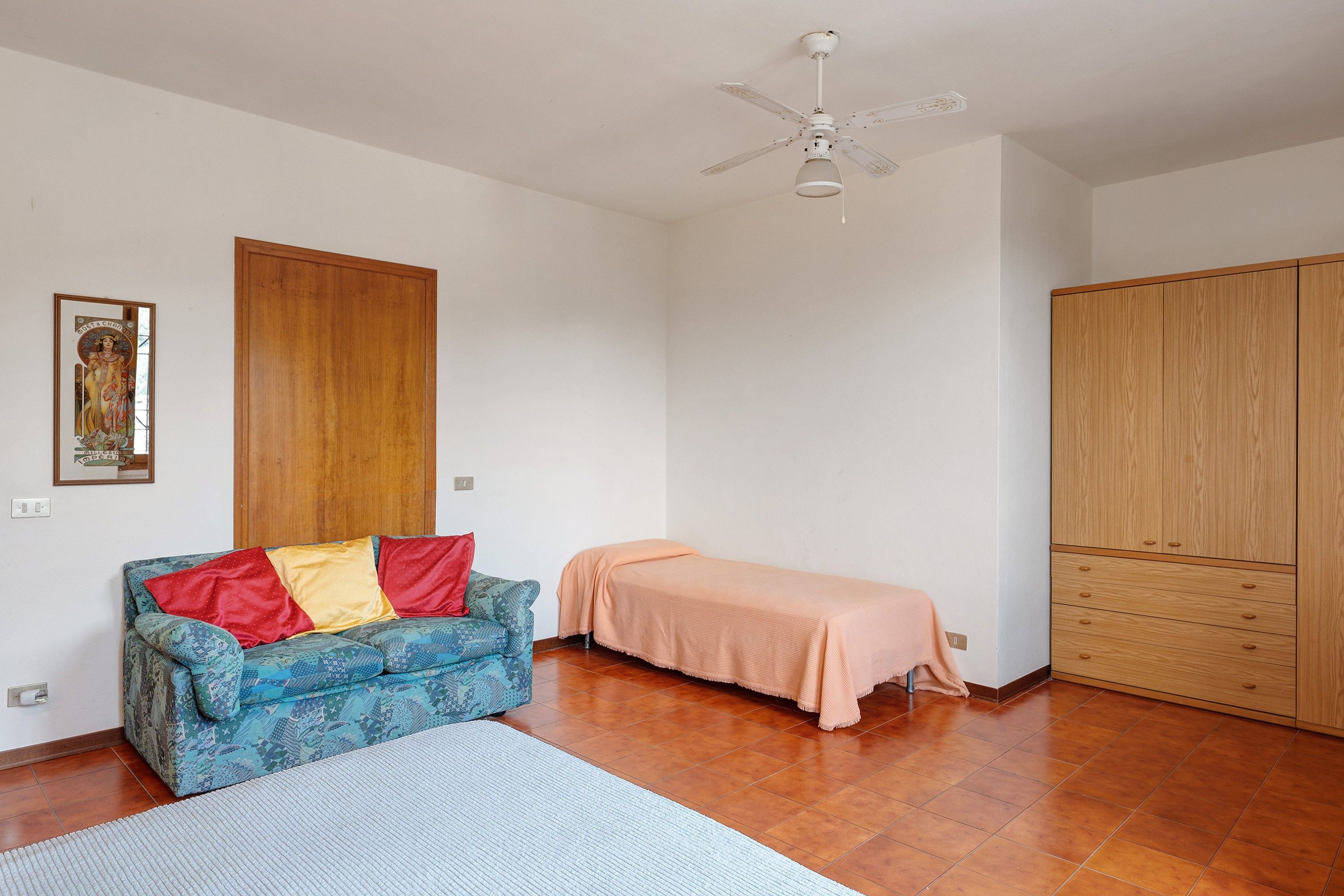 Una stanza con un armadio in legno, un letto con coperta color pesca, un divano blu con motivi e cuscini rossi e gialli, e una ventola a soffitto.