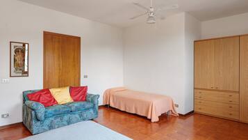 Una stanza con un armadio in legno, un letto con coperta color pesca, un divano blu con motivi e cuscini rossi e gialli, e una ventola a soffitto.