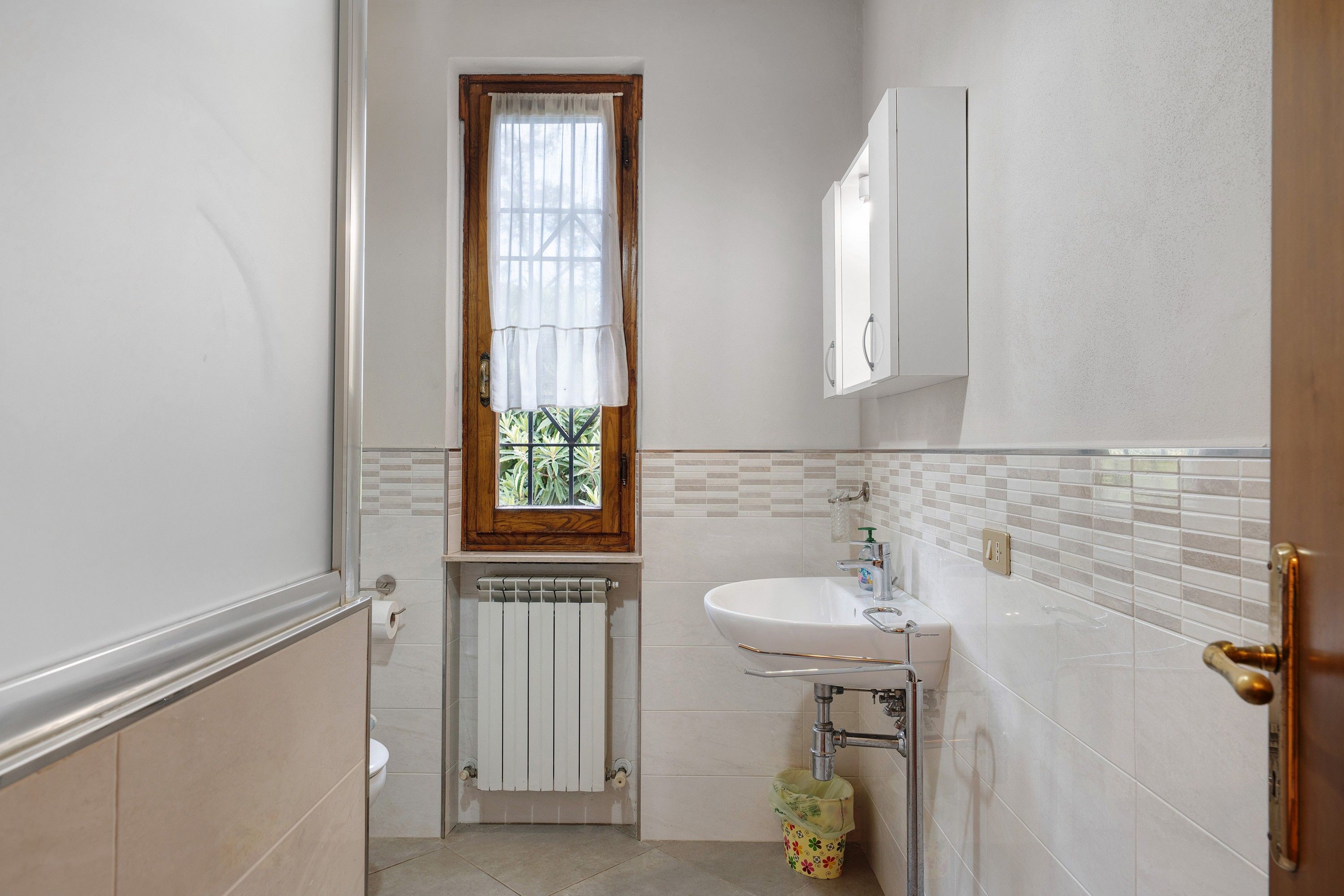 Ein Badezimmer mit Toilette, Waschbecken, einem Fenster mit Vorhang, einem Heizkörper und einem Mülleimer.