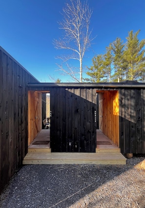 Une structure en bois dotée de deux portes ouvertes donnant sur des espaces intérieurs, un sol de gravier et un ciel bleu dégagé.
