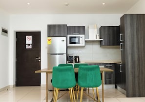 Eine moderne Küche mit einem Edelstahlkühlschrank, einer Mikrowelle und einer Kaffeemaschine. Es gibt grüne Stühle und einen Holztisch.