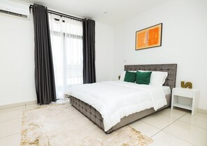 A modern bedroom with a large bed, a nightstand, a wall-mounted artwork, and a window with curtains. - Embassy Gardens Apartment by HDG (Accra)