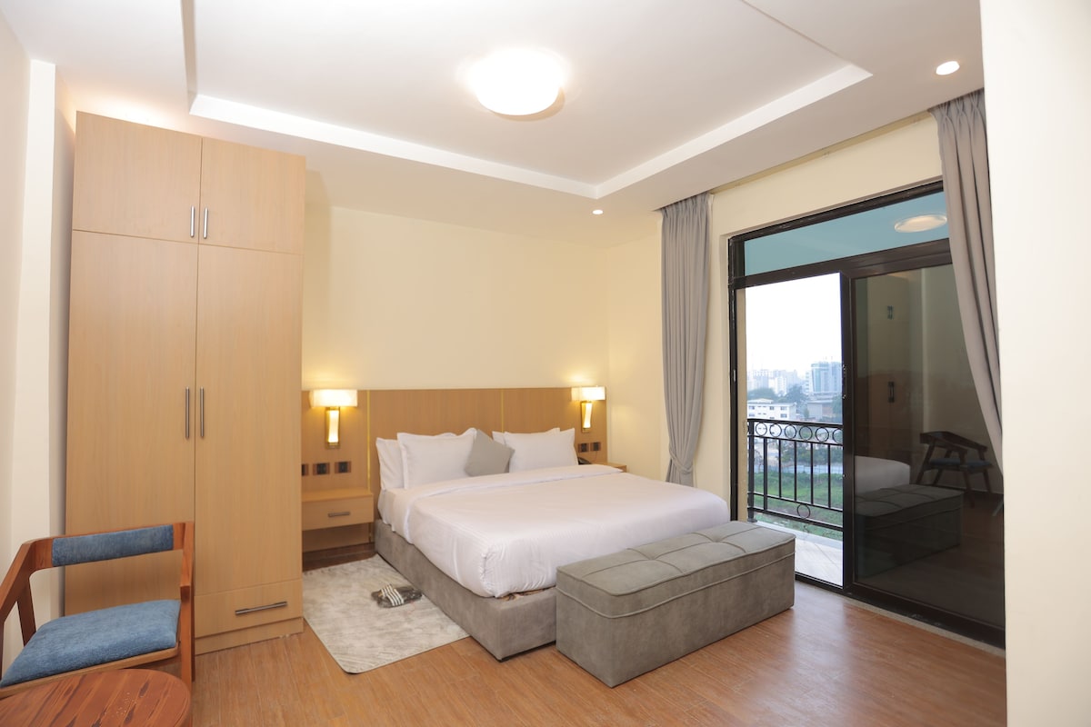 A hotel room with a bed, a wardrobe, a chair, a small ottoman, and a balcony with a view.