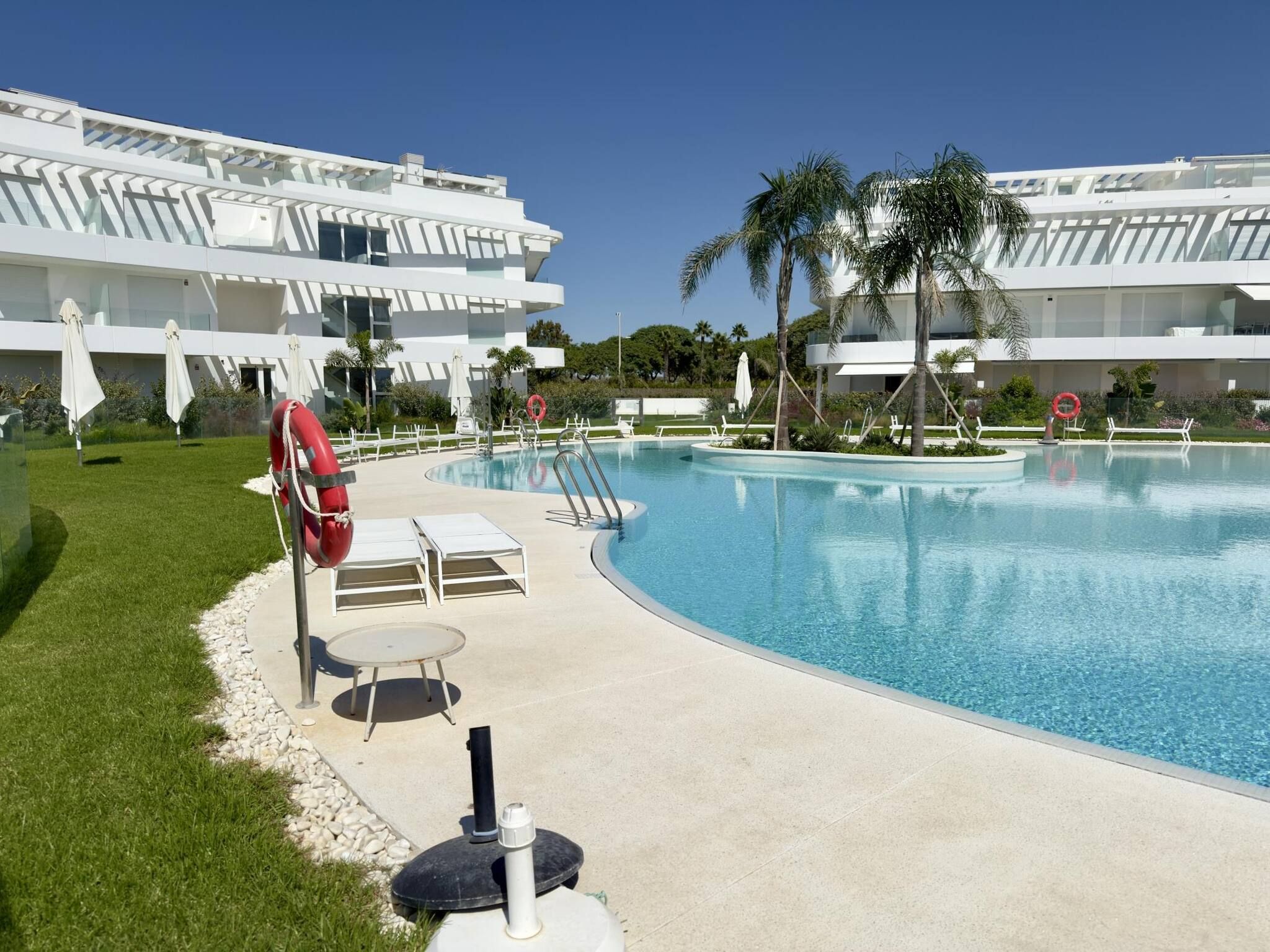 A swimming pool with lounge chairs and umbrellas, surrounded by palm trees and a modern building.