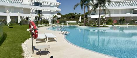 Una piscina con sillas de descanso y sombrillas, rodeada de palmeras y un edificio moderno.