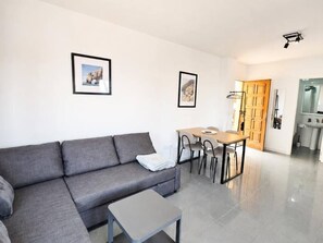 A modern living room with a grey sofa, a small table, and a dining area with chairs. There are two framed pictures on the wall. - Amber Apartment by Staycare (Rota)
