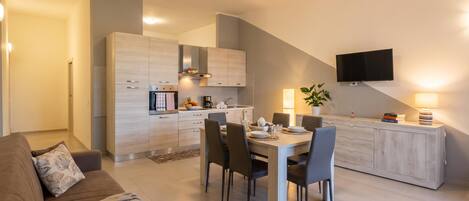 A modern kitchen with a dining area, a flat-screen TV mounted on the wall, and a sofa in the living room.