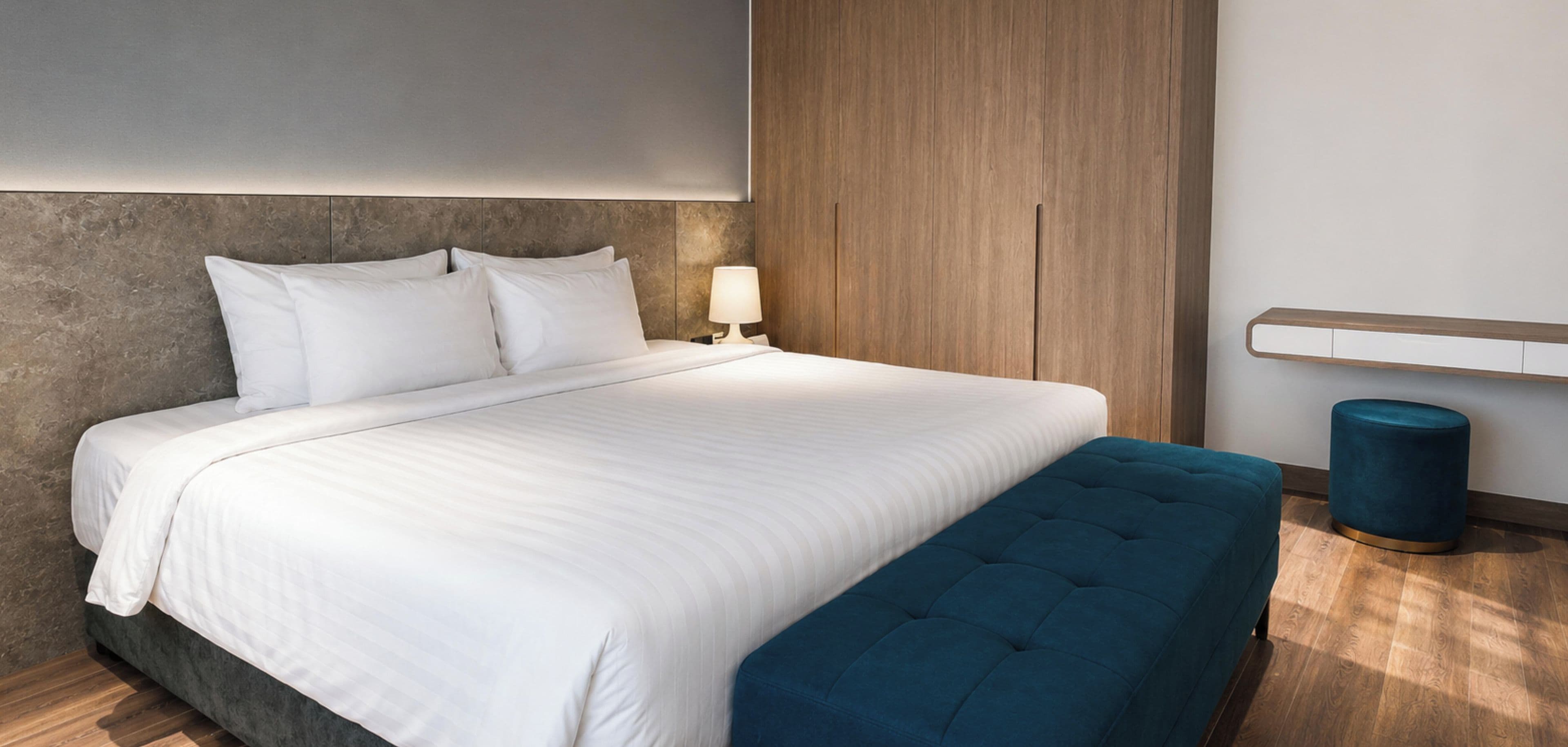 Une chambre d’hôtel moderne avec un grand lit, une table de chevet avec une lampe, un pouf bleu et un mur revêtu de panneaux en bois.