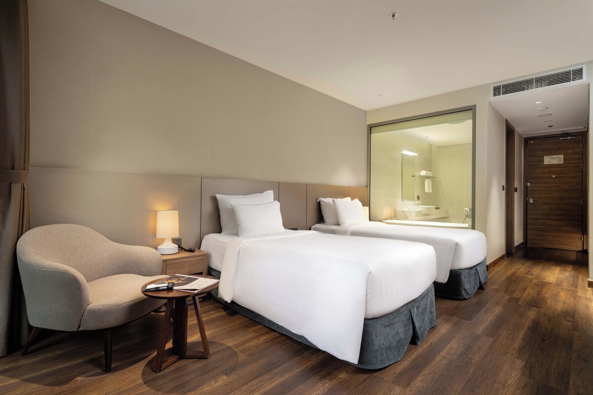 A hotel room with two beds, a chair, a nightstand, and a bathroom visible through a glass door.