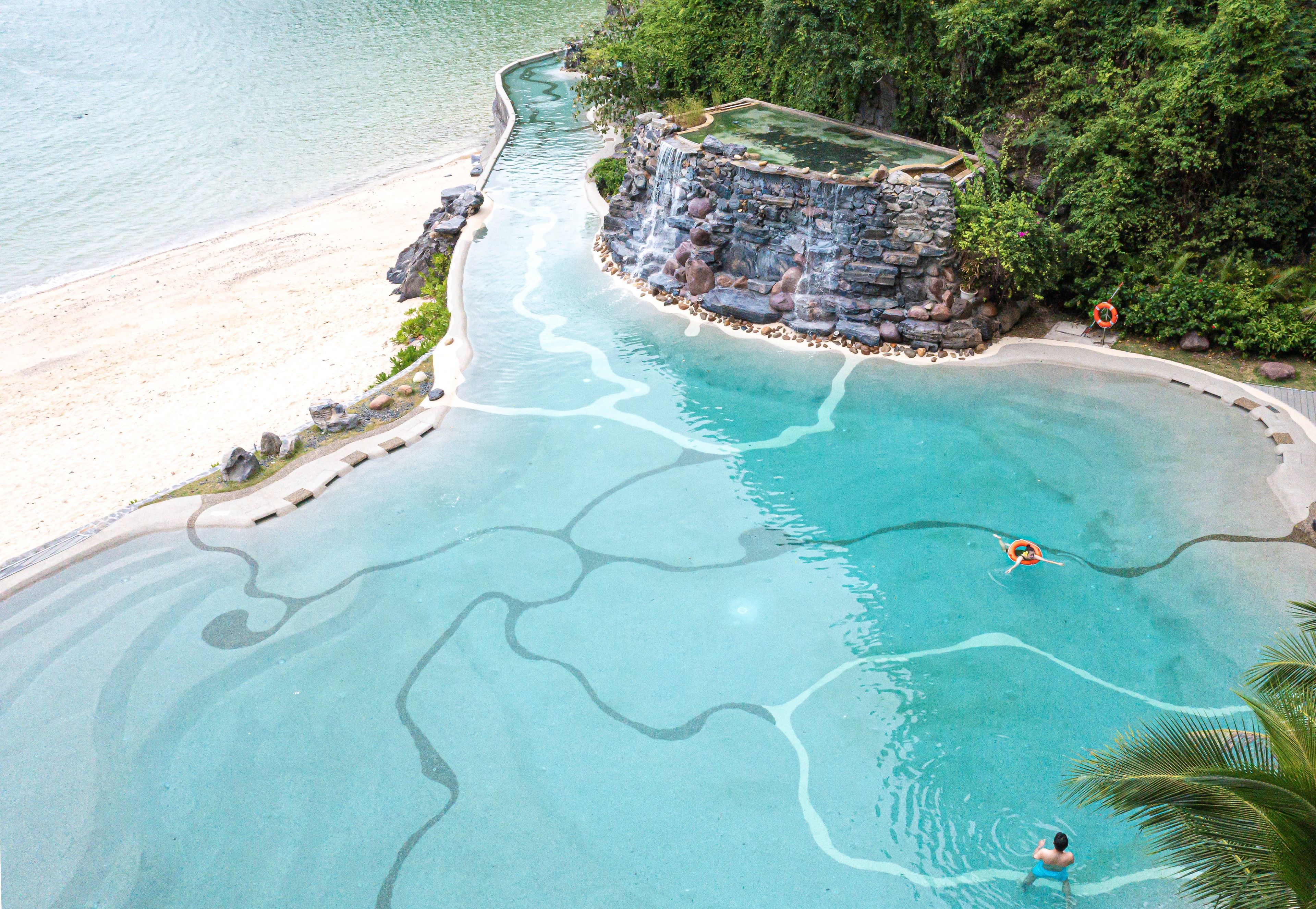 A clear blue pool with a waterfall feature, surrounded by a sandy beach and lush greenery.
