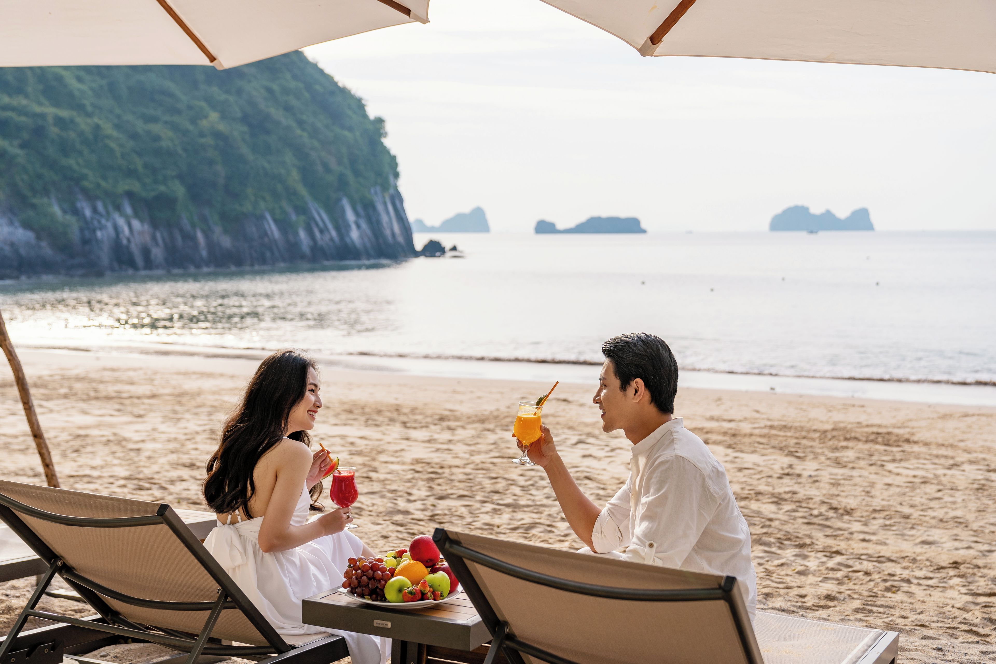 A beachside dining setup with a table, chairs, and a view of the sea and distant islands.