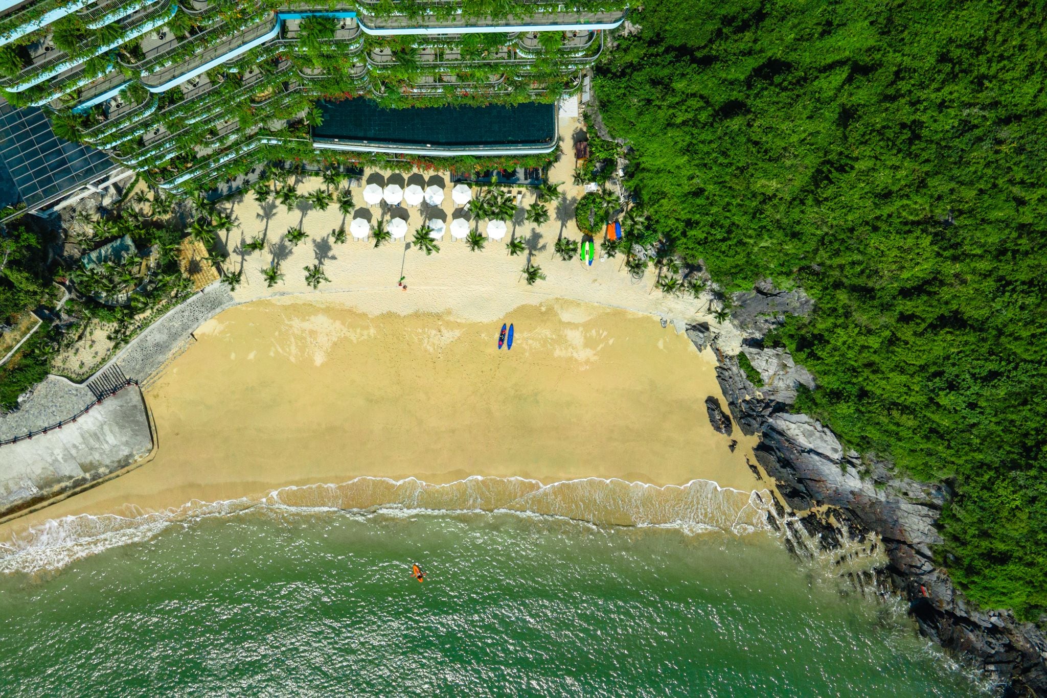 An aerial view of a beach with a pool, palm trees, and a rocky shoreline.