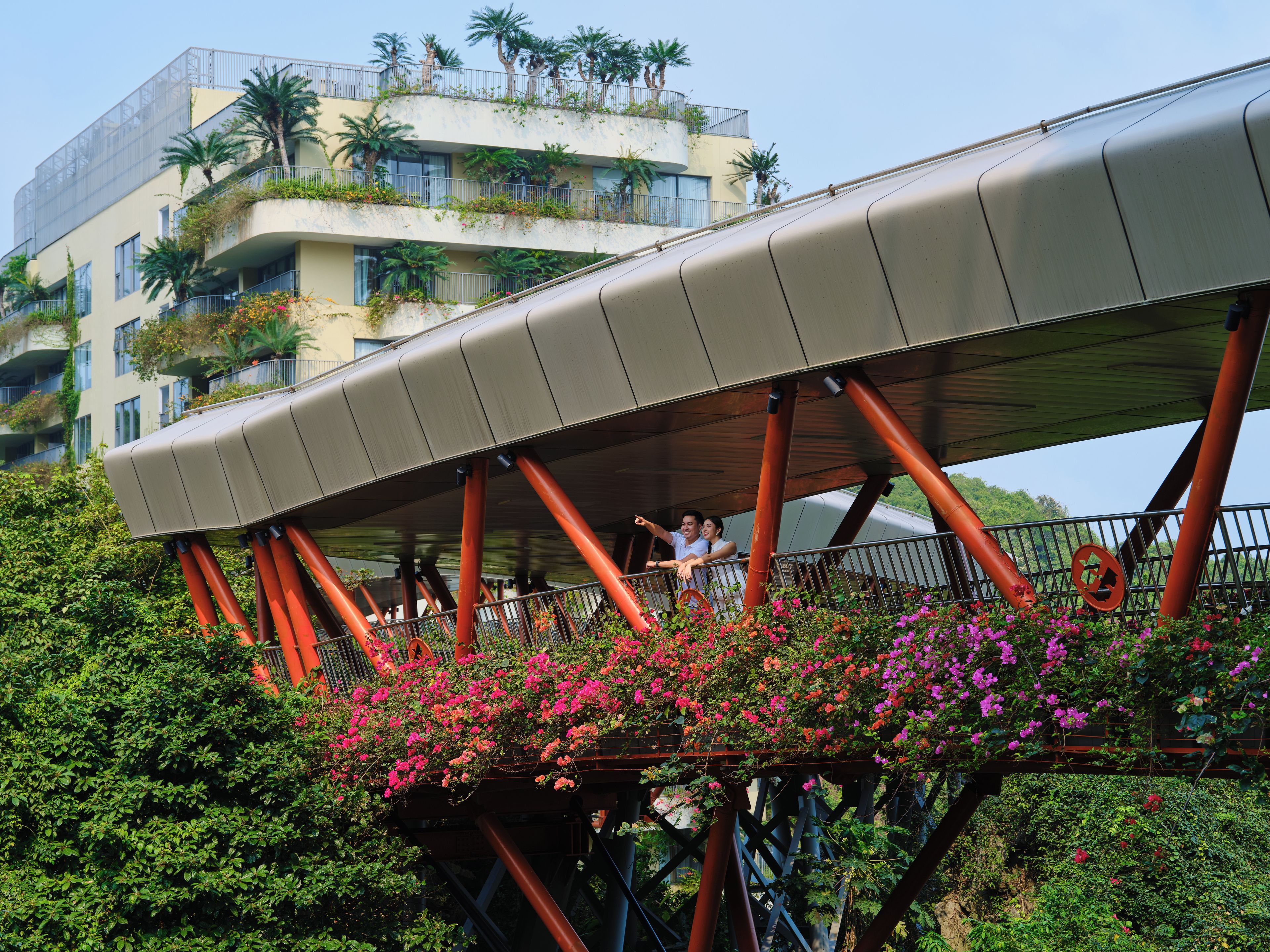 A pedestrian bridge with a curved roof supported by red pillars, surrounded by lush greenery and flowering plants.