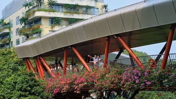 A pedestrian bridge with a curved roof supported by red pillars, surrounded by lush greenery and flowering plants.