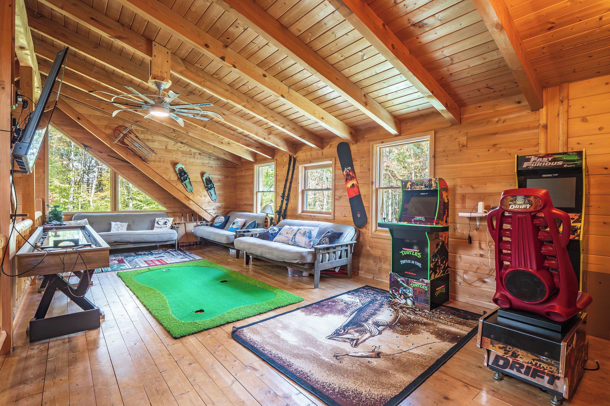 A wooden cabin interior with a green carpet, two sofas, a gaming table, and a red arcade machine.