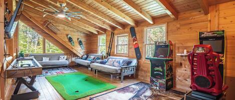A wooden cabin interior with a green carpet, two sofas, a gaming table, and a red arcade machine.