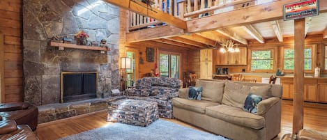 A rustic cabin interior with a stone fireplace, wooden beams, a cozy seating area, and a kitchen.