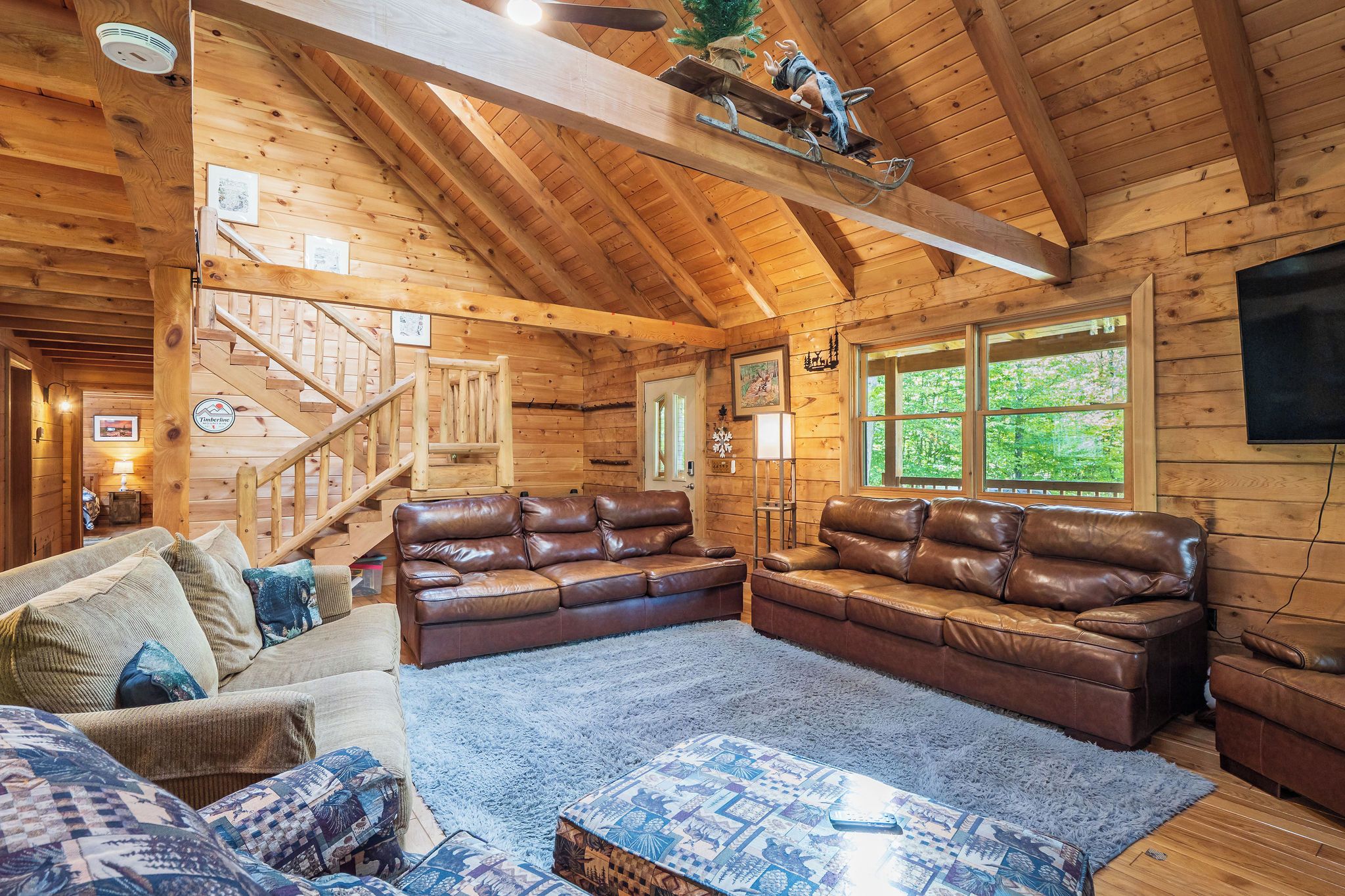 L’intérieur d’une cabane en rondins, avec un escalier en bois, des canapés en cuir, un tapis bleu et une grande fenêtre.